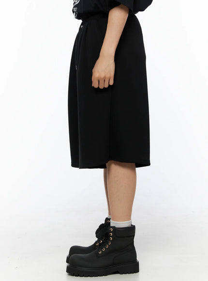 Men's Bermuda Sweat Shorts (Black) IL514 Black side shot on female model showing wide-leg profile, elastic drawstring waist, and knee-length hem paired with black lace-up boots