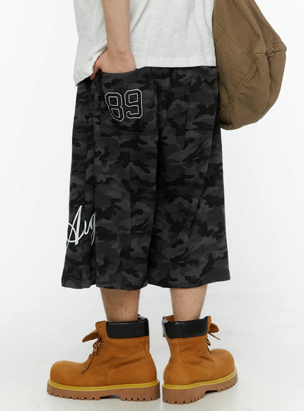 LEWKIN Men's Camo Graphic Shorts (Dark Gray) IL509 Dark gray back view on model wearing white tee and boots, hand in pocket, oversized fit with script graphic