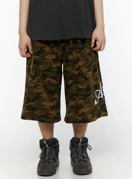 LEWKIN Men's Camo Graphic Shorts (Dark Green) IL509 front close-up on male model wearing dark gray sleeveless tee and chunky sneakers, wide-leg silhouette with script graphic