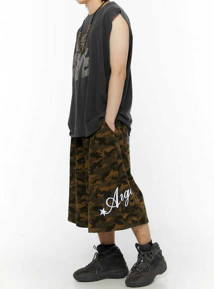 Men's Camo Graphic Shorts (Dark Green) IL509 side full-body view on male model styled with gray sleeveless graphic tee and beaded necklace, showing Angel script print and length