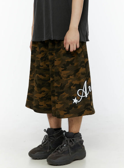 Men's Camo Graphic Shorts (Dark Green) IL509 side close-up on male model highlighting wide-leg cut, Angel script logo print and star detail on left leg, cotton fabric texture