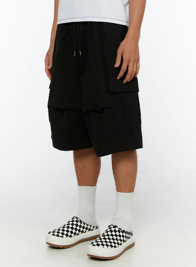 Men's Cargo Bermuda Shorts IL513 Black close-up side view on male model showing layered cargo pocket detail and wide-leg silhouette with checkered sneakers
