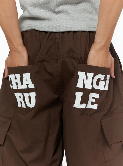 LEWKIN Men's Cargo Pocket Jorts IL510 Dark brown close-up back detail showing white CHANGE RULE graphic print on rear pockets with elastic waistband and cargo pockets visible
