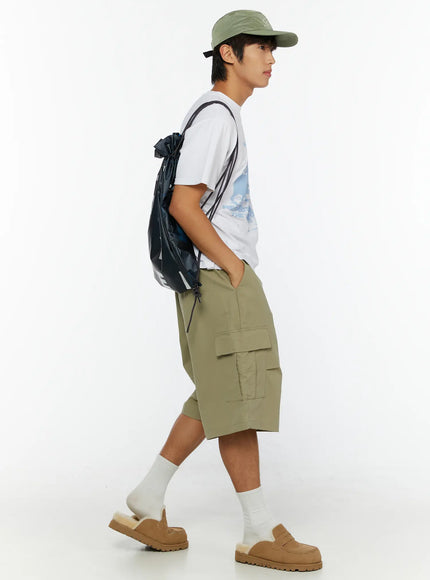 Men's Cargo Shorts IL513 Dark green side full-body view on male model walking, green cap and black drawstring backpack, cargo pocket detail visible