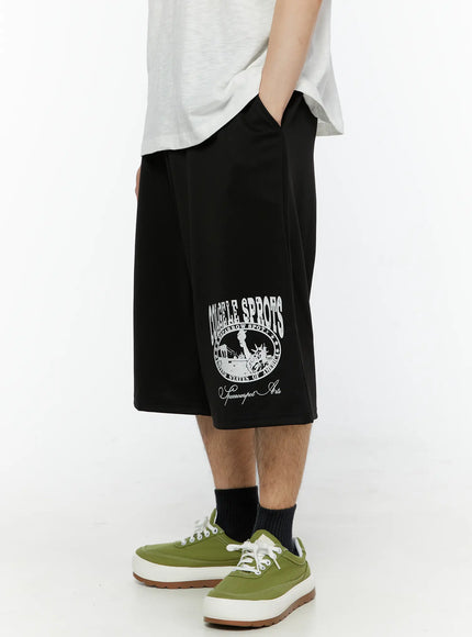 Men's Casual Number Print Shorts (Black) IL509 Black side view on model with hand in pocket, showing collegiate circle logo print and relaxed wide-leg fit with green sneakers