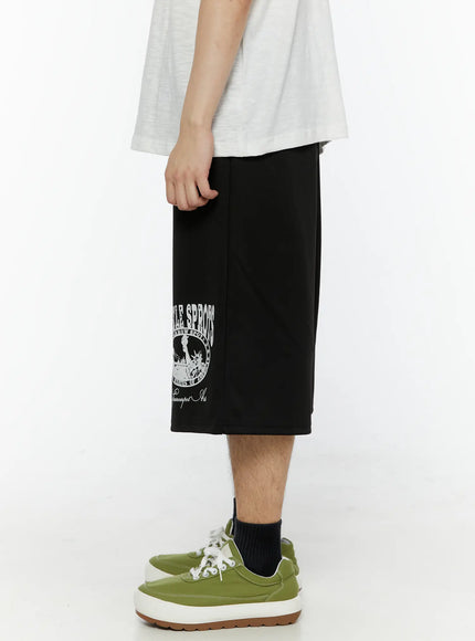 Men's Casual Number Print Shorts (Black) IL509 Black side view on model showing clean silhouette of wide-leg shorts with collegiate circle graphic partially visible, green sneakers