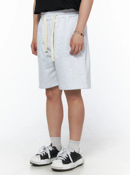 Men's Casual Sweat Shorts (Light Gray) IL502 Light Gray side view on model showing patch pocket detail, cream drawstring, elastic waist, heathered fleece texture, styled with black tee