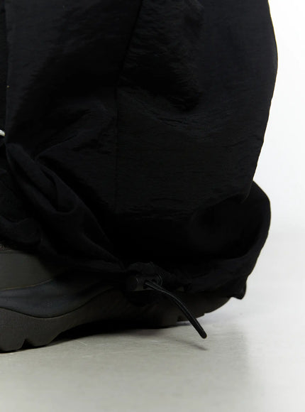 Men's Contrast Piping Nylon Track Pants (Black) IL507 Black close-up detail of hem drawstring toggle and bunched nylon fabric at ankle on model