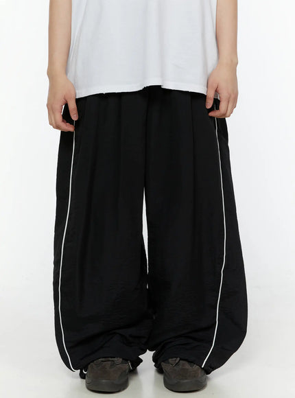 LEWKIN Men's Contrast Piping Nylon Track Pants (Black) IL507 Black S(F1) front close-up on model pulling waistband, wide-leg silhouette with white side piping