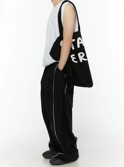 Men's Contrast Piping Nylon Track Pants (Black) IL507 Black side view on model carrying tote bag, white contrast piping detail along outer seam, gathered hem