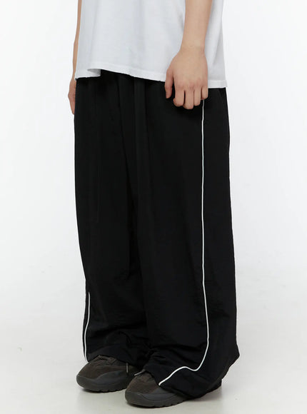 Men's Contrast Piping Nylon Track Pants (Black) IL507 Black side close-up on model showing white piping seam detail and oversized wide-leg nylon fabric drape