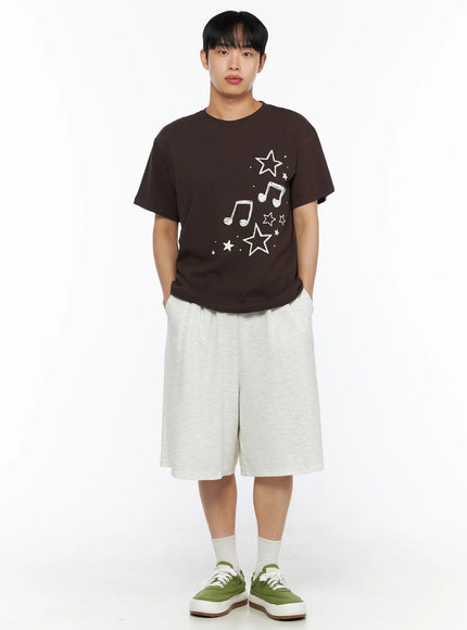 Men's Drawing Graphic Tee IL514 Brown front shot on male model hands in pockets, star and music note print, styled with grey shorts and green sneakers