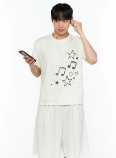 LEWKIN Men's Drawing Graphic Tee IL514 Light yellow front shot on male model holding phone with earphones, star and music note print, size S/M