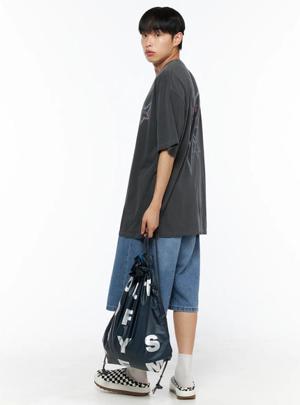 Men's Drawstring String Bag IL510 Navy side view on male model in charcoal tee holding bag by drawstring cords at side in denim shorts and checkerboard shoes