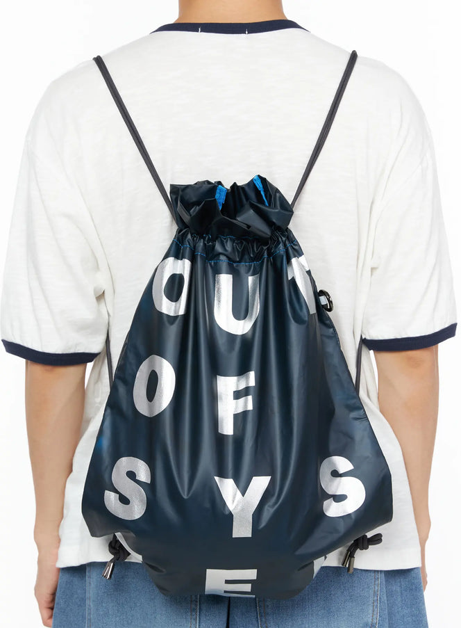 Men's Drawstring String Bag IL510 Navy back view worn on male model in white tee and jeans showing silver letter graphic and blue accent stitching