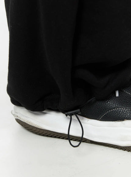 Men's Flame Wide Sweatpants (Black) IL514 close-up detail of black hem drawstring toggle at ankle cuff showing adjustable cinch mechanism on fleece fabric