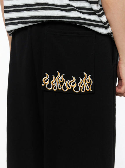LEWKIN Men's Flame Wide Sweatpants (Black) IL514 close-up detail of gold and white flame embroidery on back pocket, worn on model with striped tee