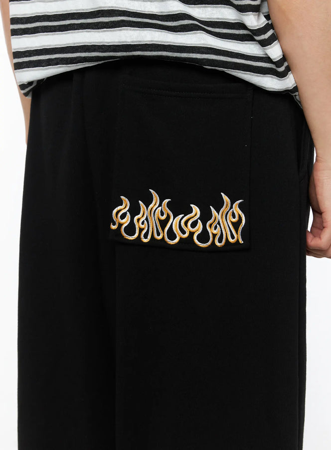 LEWKIN Men's Flame Wide Sweatpants (Black) IL514 close-up detail of gold and white flame embroidery on back pocket, worn on model with striped tee