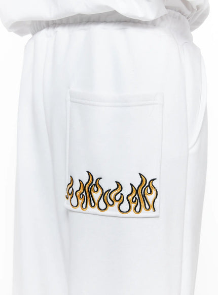 Men's Flame Wide Sweatpants (White) IL514 White close-up detail of back pocket with gold and black embroidered flame graphic on white fleece fabric, elastic waist visible
