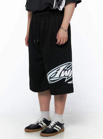 Men's Graphic Logo Shorts (Black) IL502 black close-up front view on model highlighting large blue and white graphic logo print on right leg with drawstring waist detail