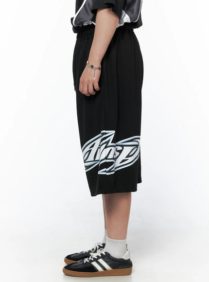 Men's Graphic Logo Shorts (Black) IL502 black side view on model cropped waist-down showing full logo graphic print on leg hem elastic waistband and wide-leg silhouette