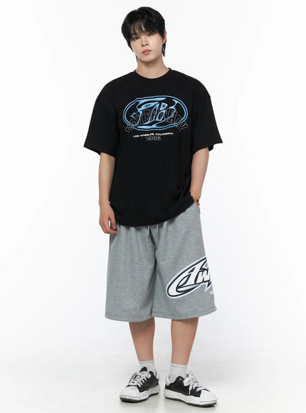 Men's Graphic Logo Shorts (Gray) IL502 Gray front view on male model in full outfit with black graphic tee, wide-leg shorts with large logo print, sneakers, standing pose