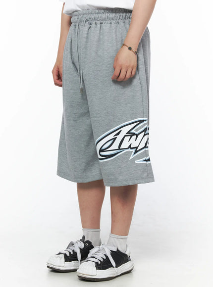 Men's Graphic Logo Shorts (Gray) IL502 Gray angled front-side view on model highlighting large blue-outlined graphic logo print detail on left leg, elastic waistband