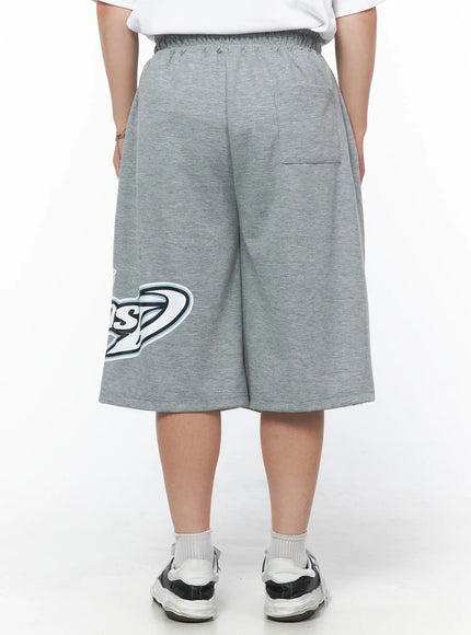 Men's Graphic Logo Shorts (Gray) IL502 Gray back view on model showing elastic waistband, back patch pocket, wide-leg silhouette, partial logo visible at lower leg