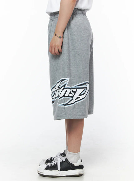 Men's Graphic Logo Shorts (Gray) IL502 Gray side view on model showing full graphic logo print across hem, wide-leg silhouette, paired with white tee and sneakers