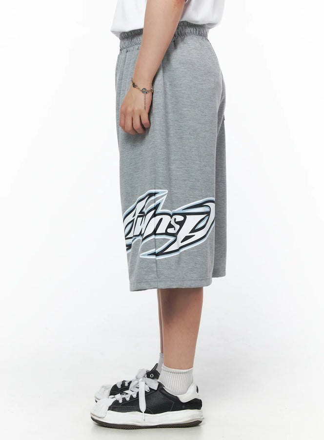 Men's Graphic Logo Shorts (Gray) IL502 Gray side view on model showing full graphic logo print across hem, wide-leg silhouette, paired with white tee and sneakers