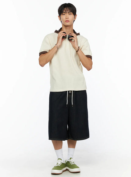 Men's Graphic Loose-Fit Shorts IL515 Dark blue full-length front view on male model wearing cream hooded polo holding headphones, wide-leg denim with drawstring and green sneakers