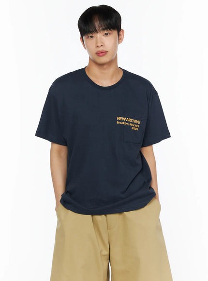 Men's Graphic Pocket Tee IL514 Navy cropped front view on male model showing yellow NEW ARCHIVE Brooklyn New York ©2018 chest pocket graphic, hands in pockets