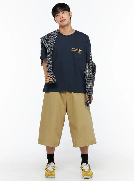 Men's Graphic Pocket Tee IL514 Navy full-body front view on male model holding open plaid shirt over shoulders, wide khaki shorts and yellow sneakers, layered street look