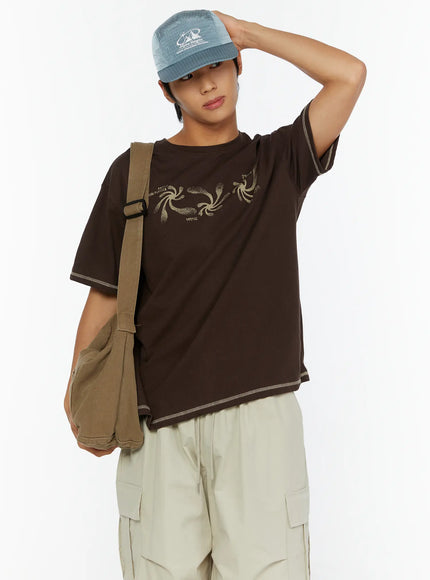 Men's Graphic Print T-Shirt IL513 Dark brown front view on male model one hand raised to cap styled with olive crossbody bag and beige cargo pants