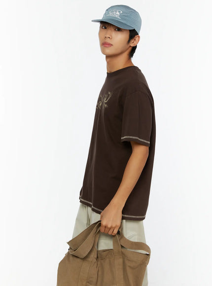 Men's Graphic Print T-Shirt IL513 Dark brown side view on male model wearing blue cap holding olive canvas bag showing contrast stitching at hem