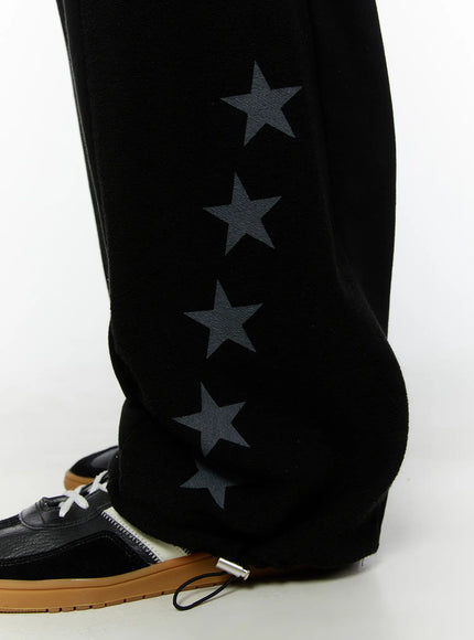 Men's Graphic Sweatpants (Black) IU529 Black close-up detail of lower leg, showing four charcoal star prints on fleece-textured fabric with wide hem opening