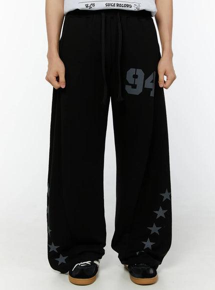 LEWKIN Men's Graphic Sweatpants (Black) IU529 Black M/L front view on model, wide-leg fleece trousers with '94' graphic print and star motifs at hem, drawstring waist