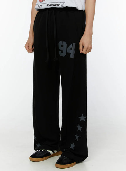 Men's Graphic Sweatpants (Black) IU529 Black slightly angled front view on model, wide-leg sweatpants with '94' number print on thigh and graduating star prints near hem