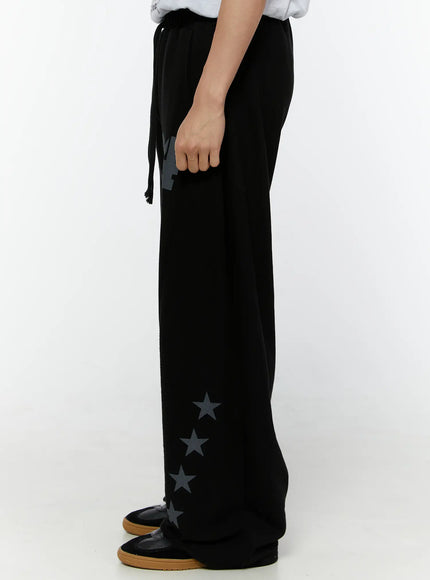Men's Graphic Sweatpants (Black) IU529 Black side view on model, showing wide-leg silhouette with drawstring waist and vertical row of star graphic prints along side hem