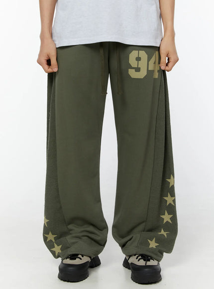 LEWKIN Men's Graphic Sweatpants (Dark Green) IU529 Dark green front view on model showing wide-leg silhouette, gold 94 print, drawstring waist, fleece side panels