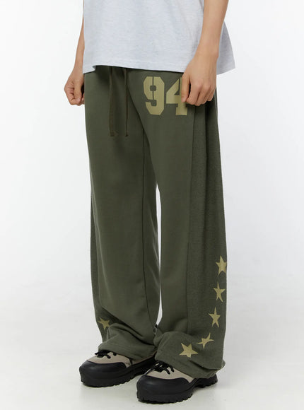 Men's Graphic Sweatpants (Dark Green) IU529 Dark green angled front view on model highlighting 94 graphic, wide ultra-baggy leg with star-print fleece side panel