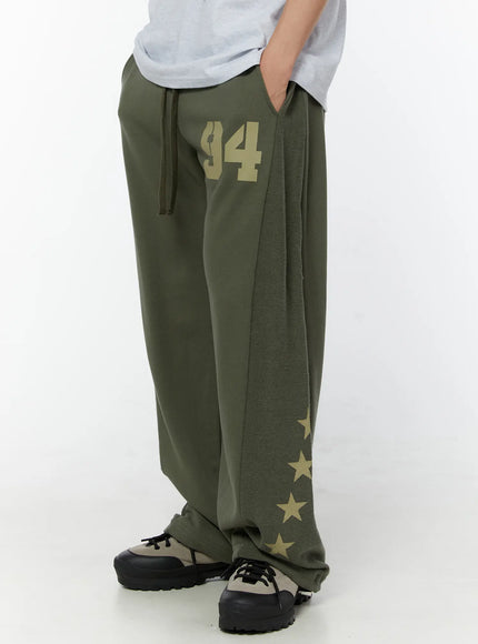 Men's Graphic Sweatpants (Dark Green) IU529 Dark green side view on model showing hand-in-pocket pose, fleece panel, gold star cluster at wide hem, and drawstring waist