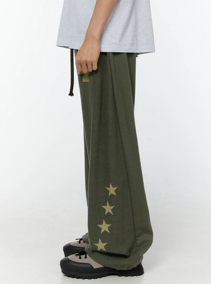 Men's Graphic Sweatpants (Dark Green) IU529 Dark green side view on model showing reverse-fleece panel, gold star print cascading down leg, wide-leg silhouette with chunky sneakers