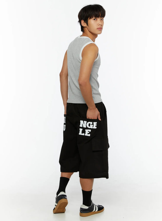 Men's Graphic Wide Jorts IL510 Black three-quarter back side view on male model in grey tank top, hand in pocket, NGLE LE print visible