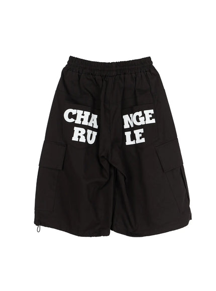 Men's Graphic Wide Jorts IL510 Black flat-lay three-quarter front angle showing CHANGE RULE graphic, cargo pockets, and hem cord toggles