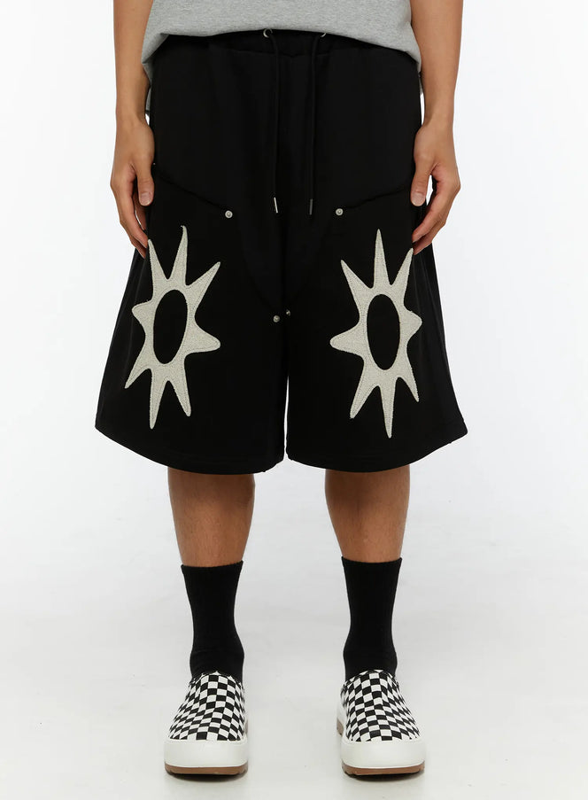 LEWKIN Men's Happy Sun Jorts IL510 Black front view on model wearing gray tee, dual cream sun appliqué patches on wide-leg shorts with drawstring waist