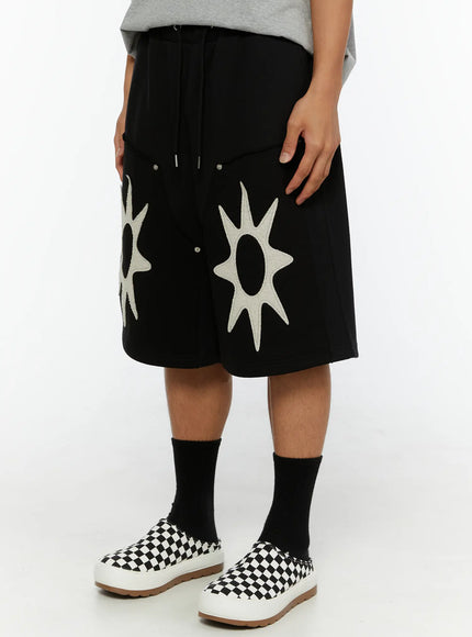 Men's Happy Sun Jorts IL510 Black angled side view on model showing cream star-burst appliqué detail, oversized silhouette with snap hardware and drawstring