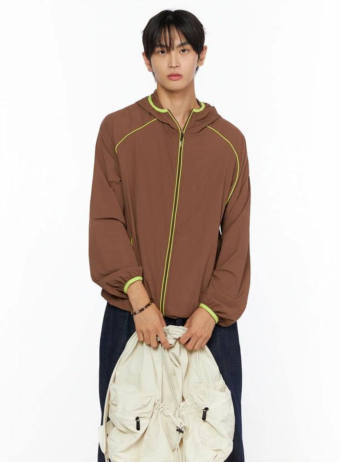 Men's Hooded Windbreaker IL528 Brown front shot on model holding cream bag, neon yellow zip and cuff trim, paired with wide-leg dark denim