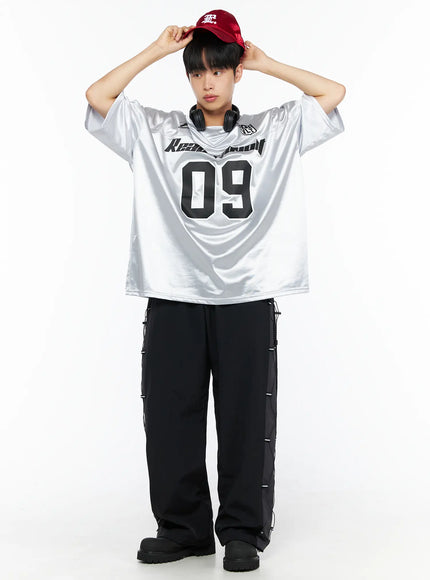 Men's Lace-Up Track Pants (Black) IL514 Black full-length front shot on male model styled with silver football jersey and red cap, adjusting cap with both hands raised