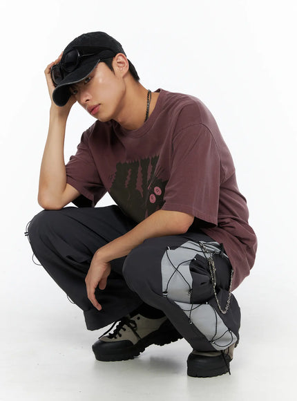 Men's Lace-Up Track Pants (Dark Gray) IL514 dark gray crouching pose shot on model in burgundy tee and black cap, hand raised to cap brim, side lace-up and chain detail visible